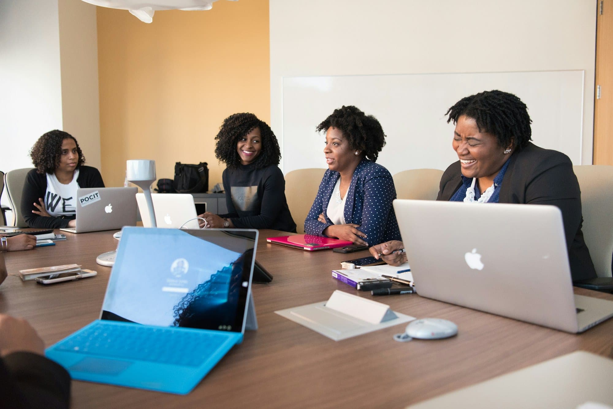 Four colleagues in discussion around a meeting table with open laptops.